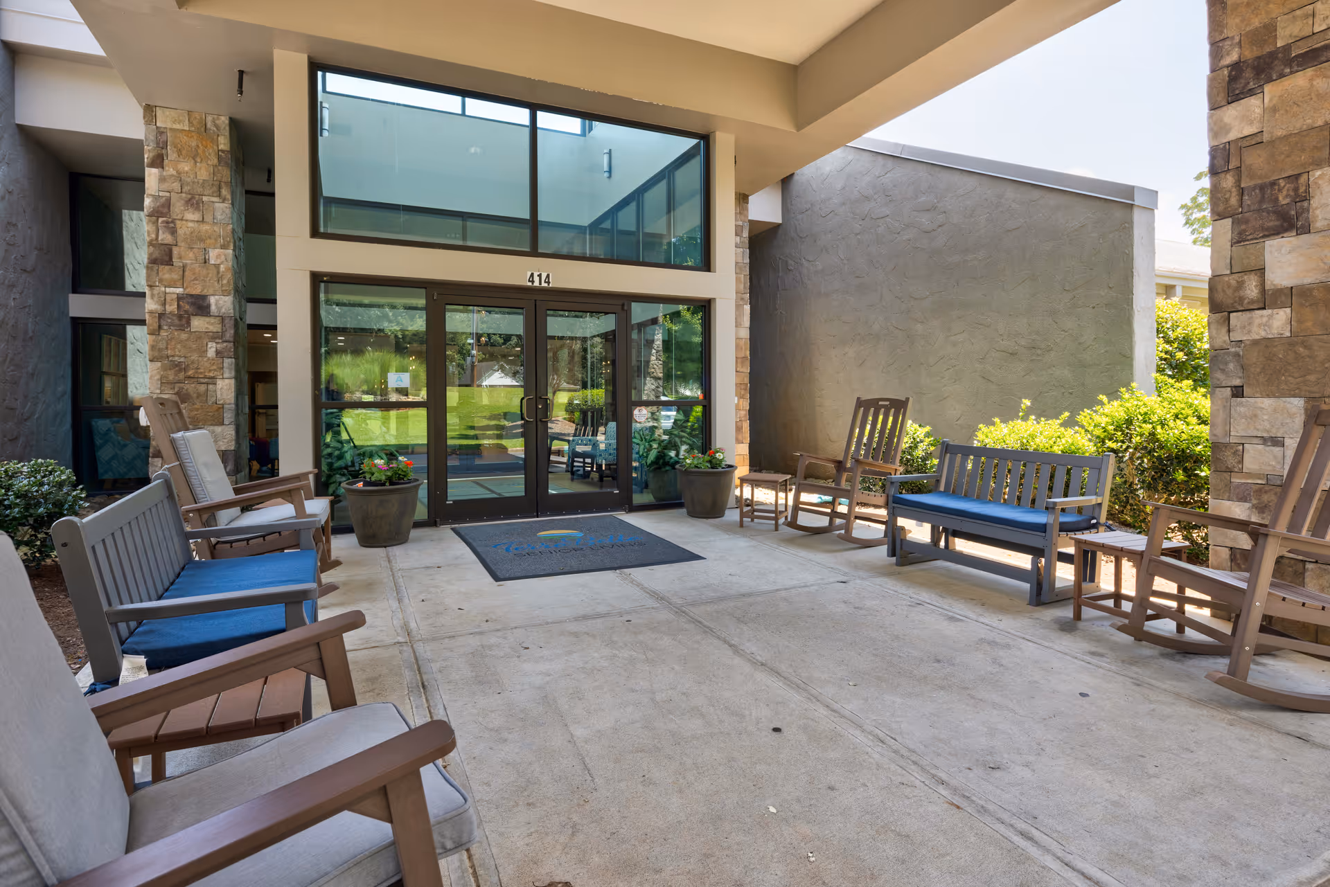 Entrance area of TerraBella Summit featuring a covered patio with multiple wooden chairs and benches with blue cushions arranged along the sides. The entrance has large glass double doors with the number 414 above them, flanked by stone pillars and potted plants.