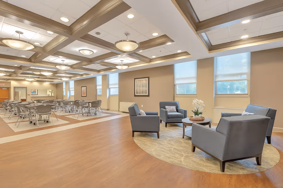 A spacious and well-lit common area in a senior living facility featuring a seating area with four gray armchairs arranged around a round coffee table with a flower arrangement. In the background, there are multiple tables and chairs set up for dining. The room has large windows with blinds, beige walls, and a ceiling with recessed lighting and decorative beams.