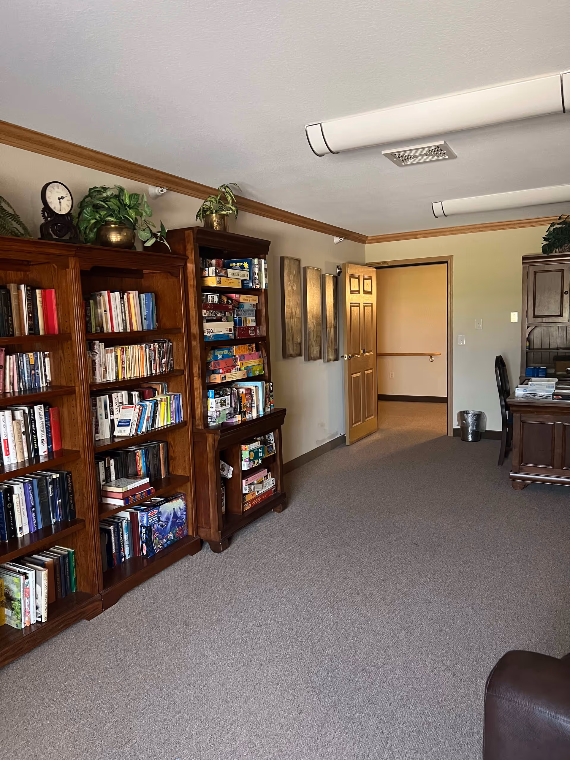 Interior room with carpeted floor featuring wooden bookshelves filled with books and board games. There is a wooden desk with a chair on the right side, and an open door leading to another room. The walls have framed artwork and the ceiling has fluorescent lights.