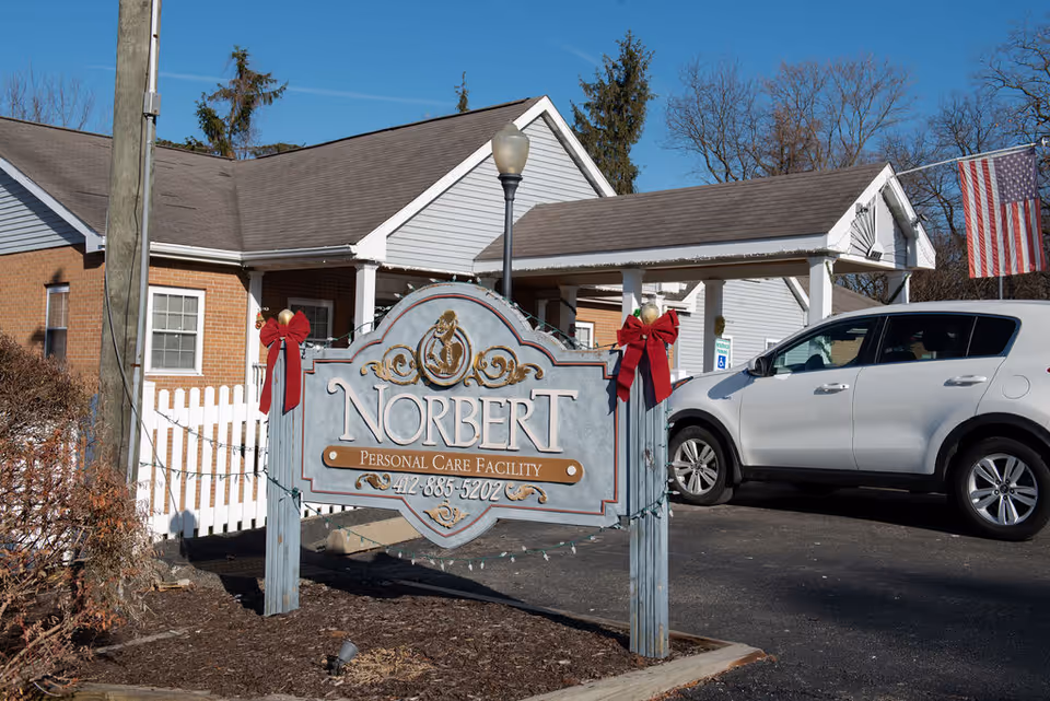 Exterior view of Norbert Personal Care Facility with a decorative sign in the foreground displaying the facility's name and phone number. The sign is adorned with red bows and holiday lights. Behind the sign is a building with a covered entrance, a white picket fence, a parked white SUV, and an American flag on a pole.