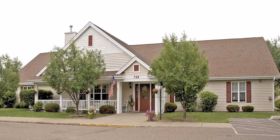 Front view of a single-story senior living facility with a porch, trees, and manicured lawn.