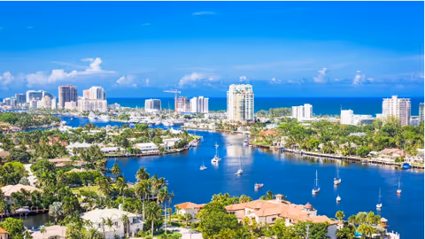 Exterior view of a coastal city with a waterway, boats, palm trees, and high-rise buildings.