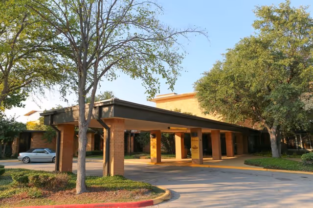 Exterior view of a senior living facility entrance with a covered drop-off area supported by brick columns, surrounded by trees and landscaping under a clear sky.