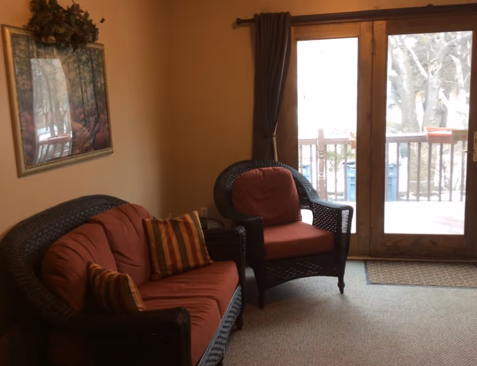 Cozy sitting area with wicker loveseat and armchair with red cushions beside glass doors leading to a deck.