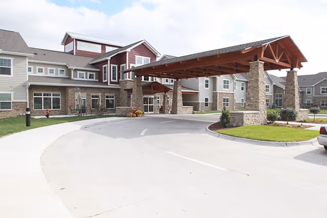 Front entrance of a multi-story senior living building with a covered porte-cochere, stone columns, and a circular driveway.