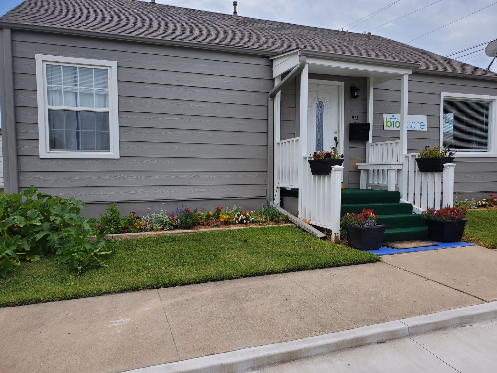 Gray single-story senior care building with white porch, green steps, and flower-filled planters.