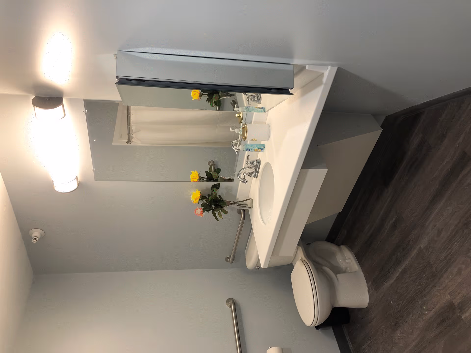 A clean bathroom with a white sink and countertop, a toilet, a large mirror above the sink, a wall-mounted light fixture, and a shower curtain visible in the reflection. There are yellow and pink flowers in a vase on the countertop, and the floor has dark wood-like flooring. Safety grab bars are installed near the toilet and sink.