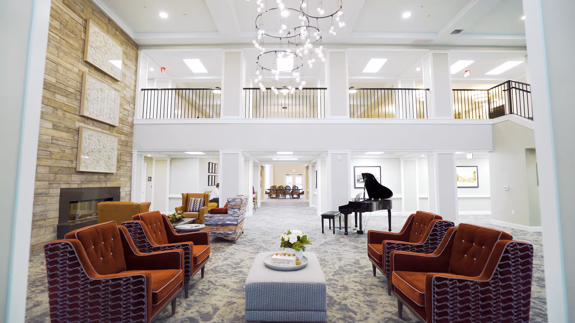 Spacious and well-lit senior living facility common area with high ceilings, modern chandeliers, and a second-floor balcony railing. The room features multiple seating arrangements with orange and patterned armchairs, a gray ottoman with a flower centerpiece, a grand piano, and a stone fireplace on the left wall. The area extends into a dining space in the background.