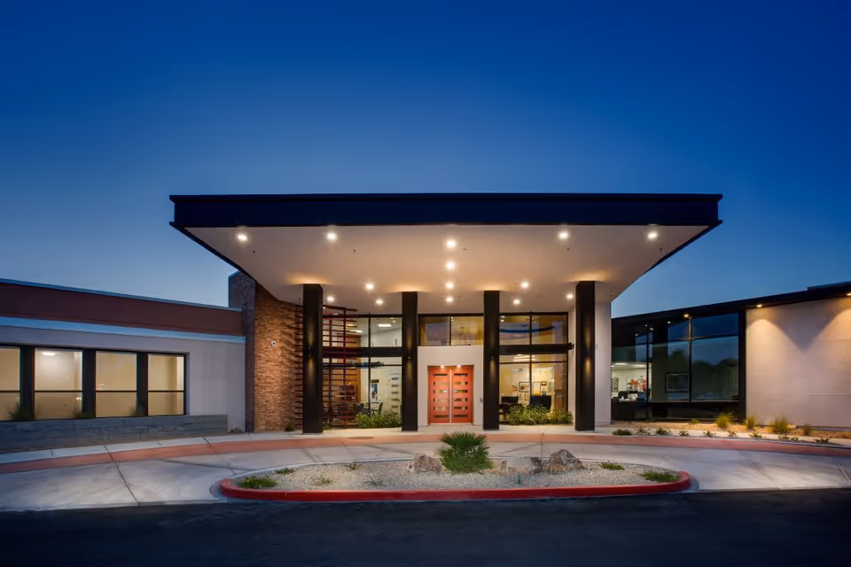 Front exterior view of Lumina Las Vegas facility at dusk with illuminated entrance featuring a covered drop-off area supported by black columns, large glass windows, and a red double door.