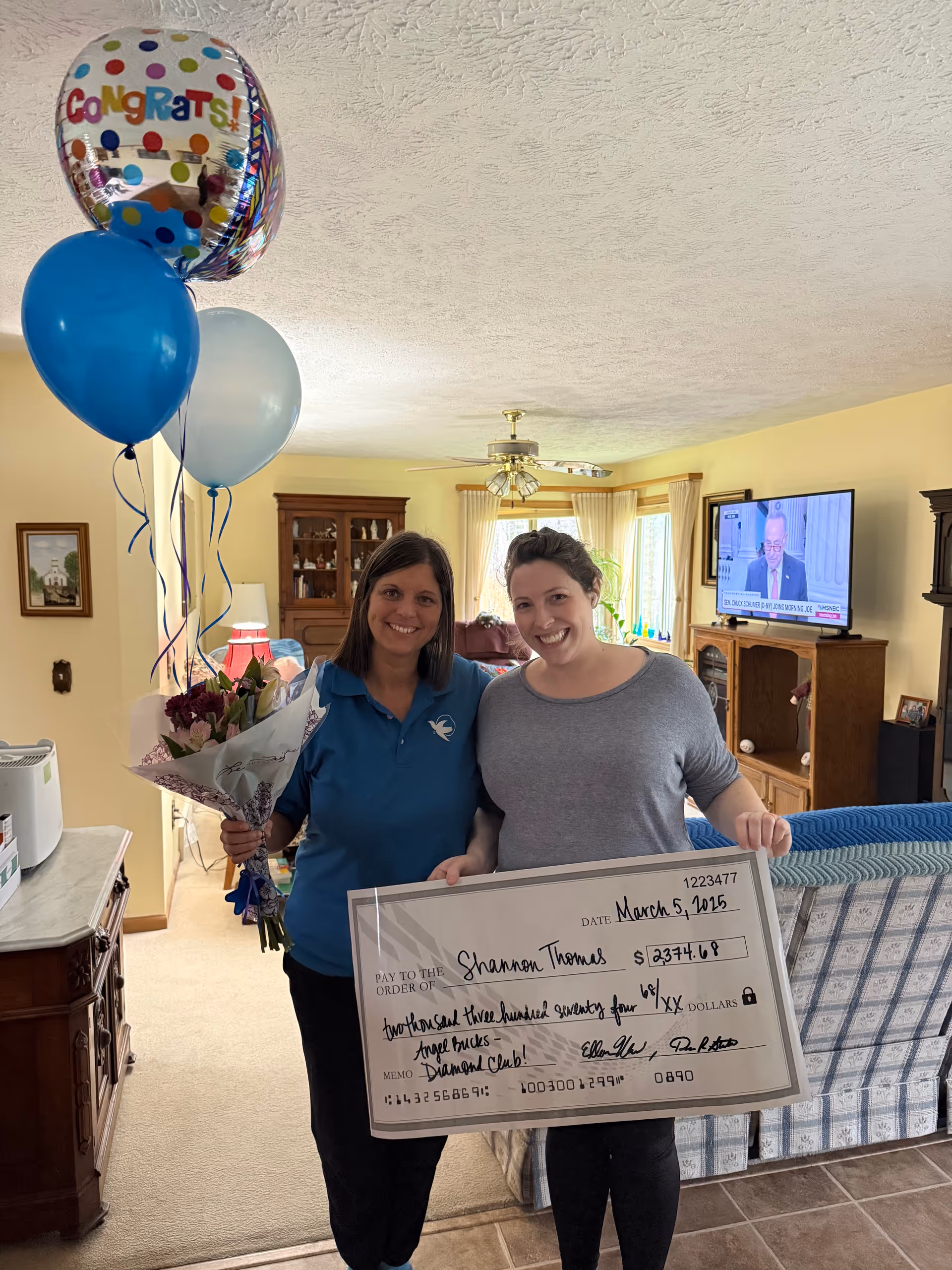 Two smiling women stand in a living room with balloons and a large check.