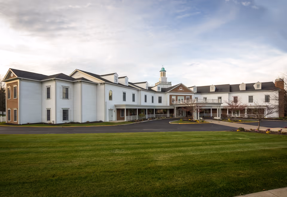 Exterior view of a large, two-story assisted living facility building with white siding and brick accents. The building features multiple windows, a covered entrance with columns, and a cupola on the roof. There is a paved driveway and a well-maintained green lawn in front of the building under a partly cloudy sky.