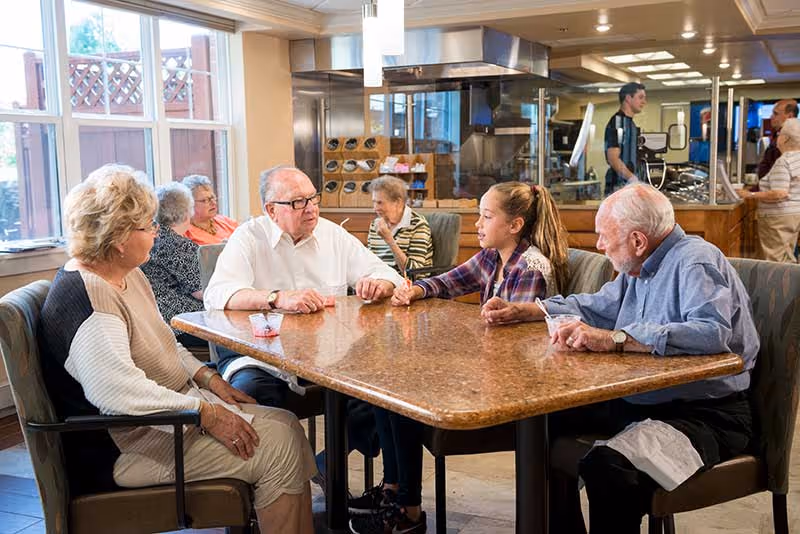 Several elderly residents and a young visitor sit and converse around a table in a senior living dining area with the kitchen and staff visible in the background.