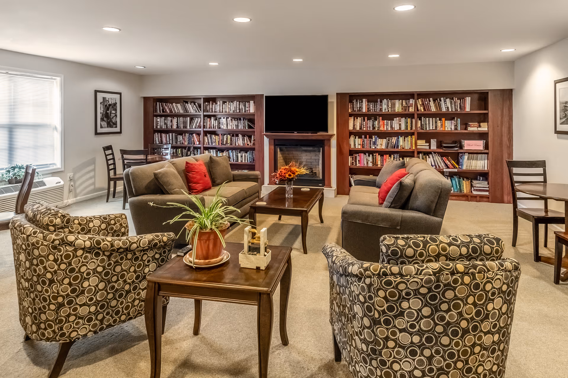 Cozy senior living room with sofas, patterned chairs, bookshelves, and a fireplace.