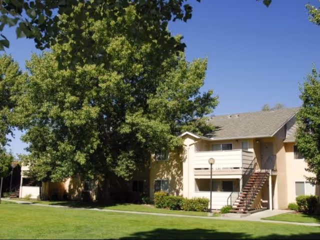 Exterior view of a two-story residential building with beige walls and a sloped roof, surrounded by green grass, bushes, and large leafy trees under a clear blue sky.