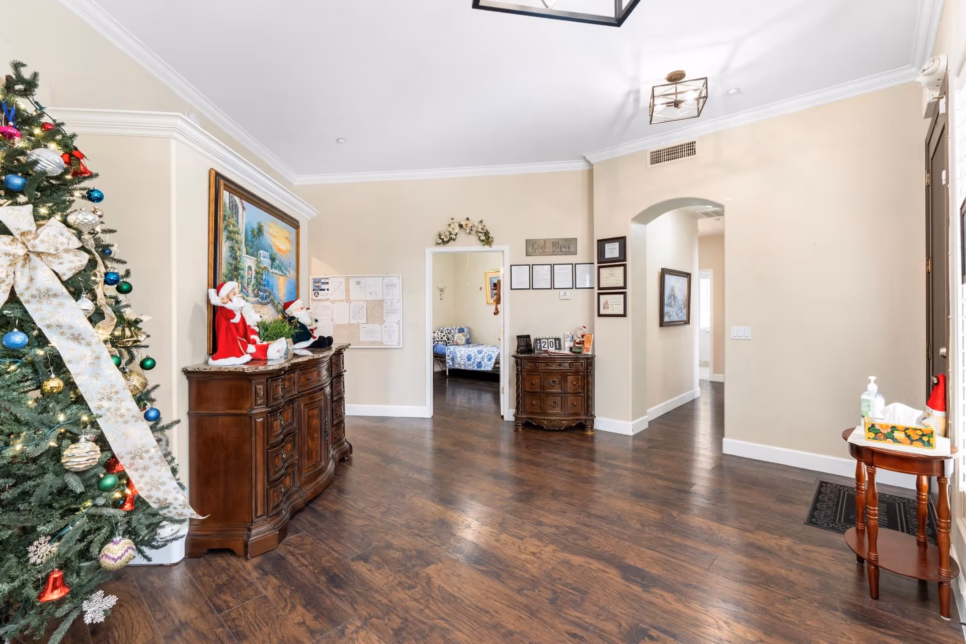 A spacious interior room with wooden flooring and beige walls, featuring a decorated Christmas tree on the left side. There are two wooden dressers with holiday decorations and framed pictures on the walls. A doorway leads to a bedroom with a blue floral bedspread. A small round table on the right holds a tissue box and hand sanitizer near the entrance door.