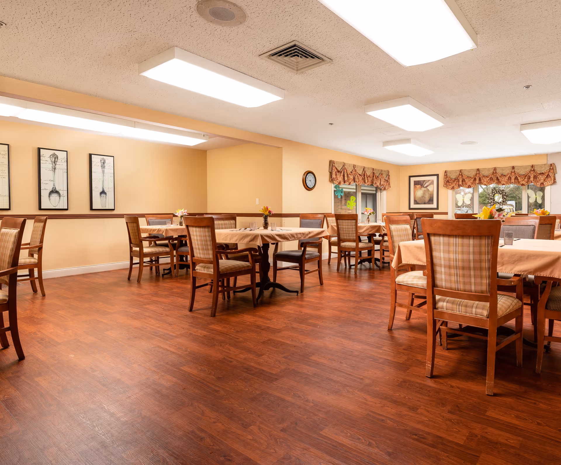 A well-lit dining room with several wooden tables covered with beige tablecloths and surrounded by wooden chairs with plaid cushions. The room has warm beige walls, framed artwork of utensils, a clock on the wall, and windows with decorative valances. Small flower arrangements are placed on the tables.