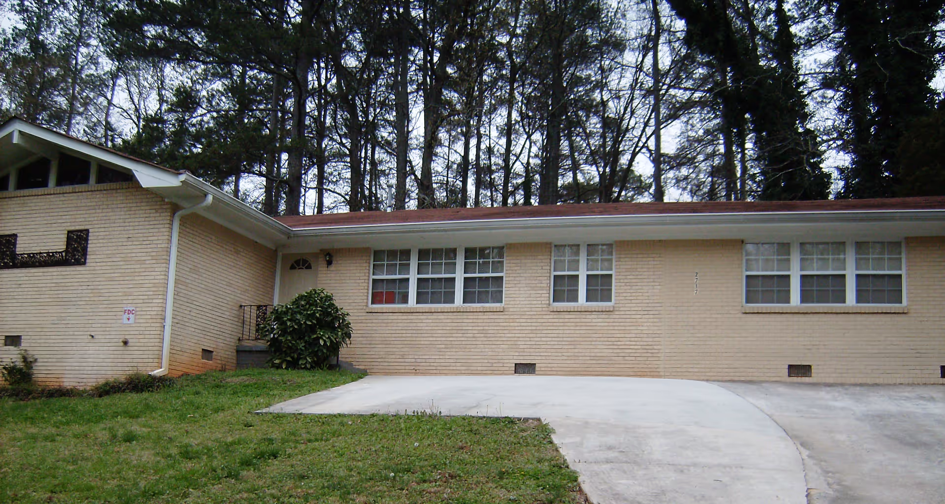Exterior view of a single-story beige brick building with multiple windows and a sloped roof, surrounded by tall trees and a grassy lawn with a concrete driveway in front.