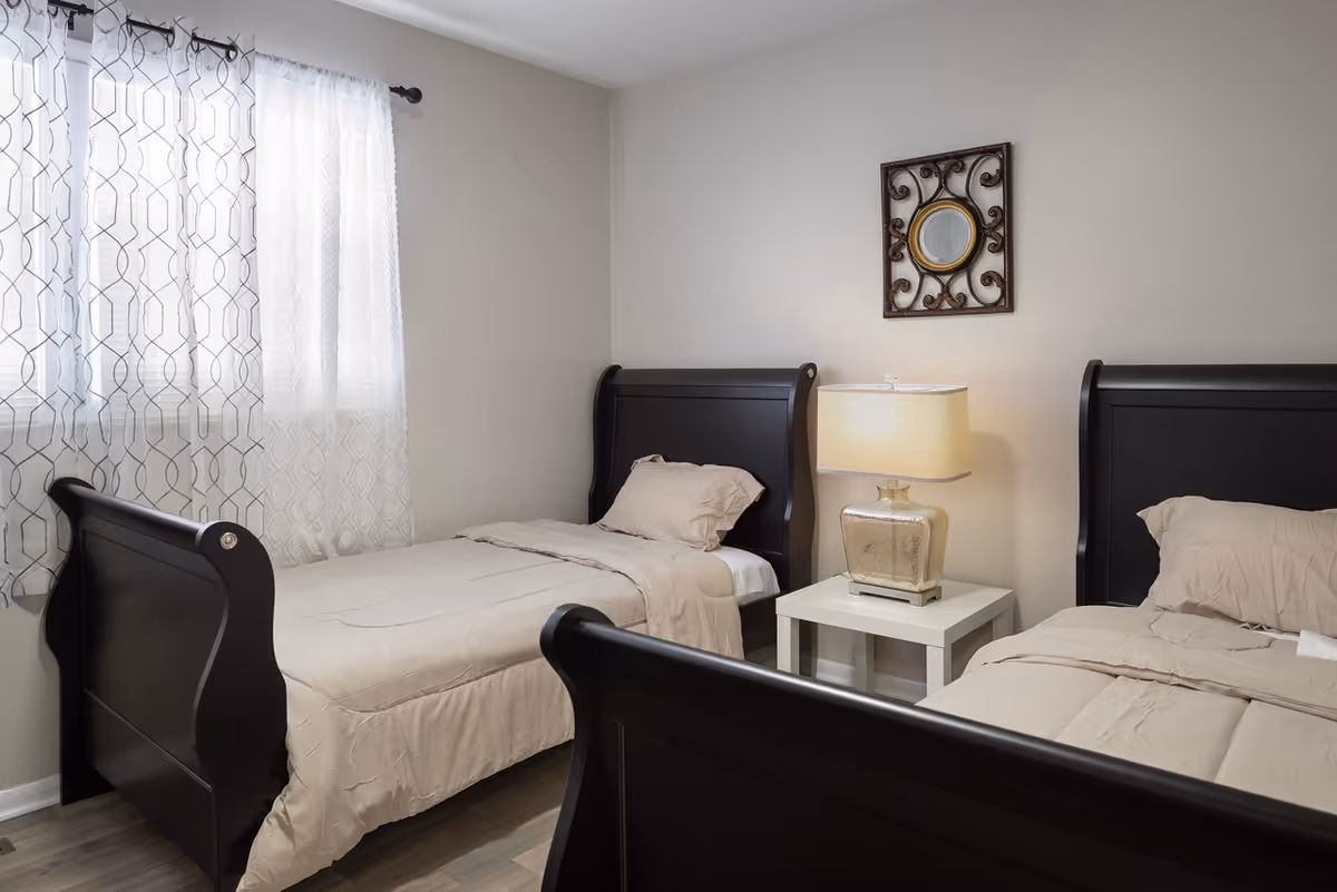 Two twin beds with beige bedding and a nightstand lamp in a neutral bedroom.
