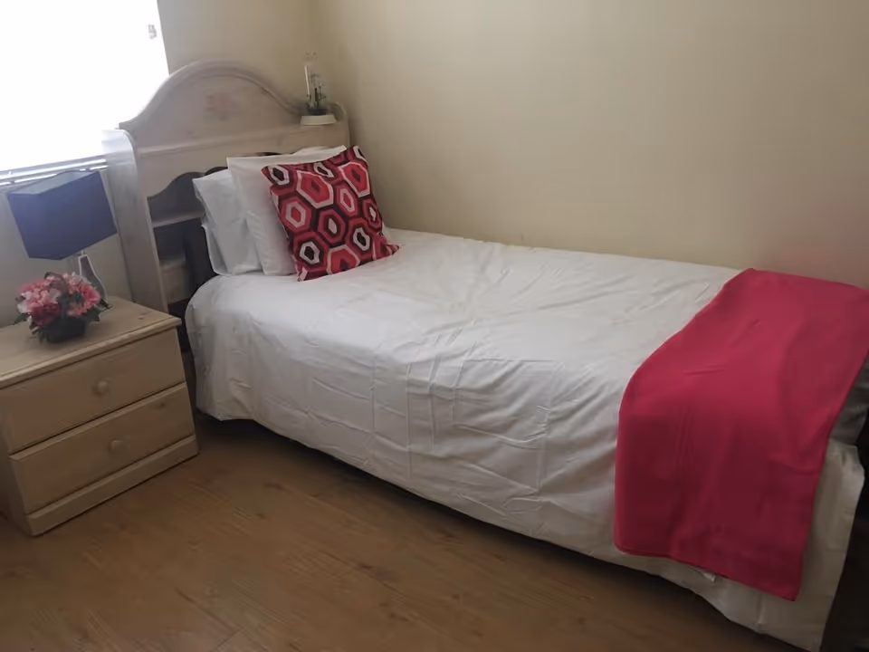A single bed with white bedding, a pink and red geometric patterned pillow, and a bright pink throw blanket at the foot of the bed. Next to the bed is a wooden nightstand with two drawers, a black lampshade, and a small flower arrangement. The room has light-colored walls and wooden flooring.