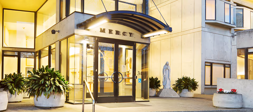 Exterior entrance of a building labeled Mercy with glass doors, planters, and a statue.