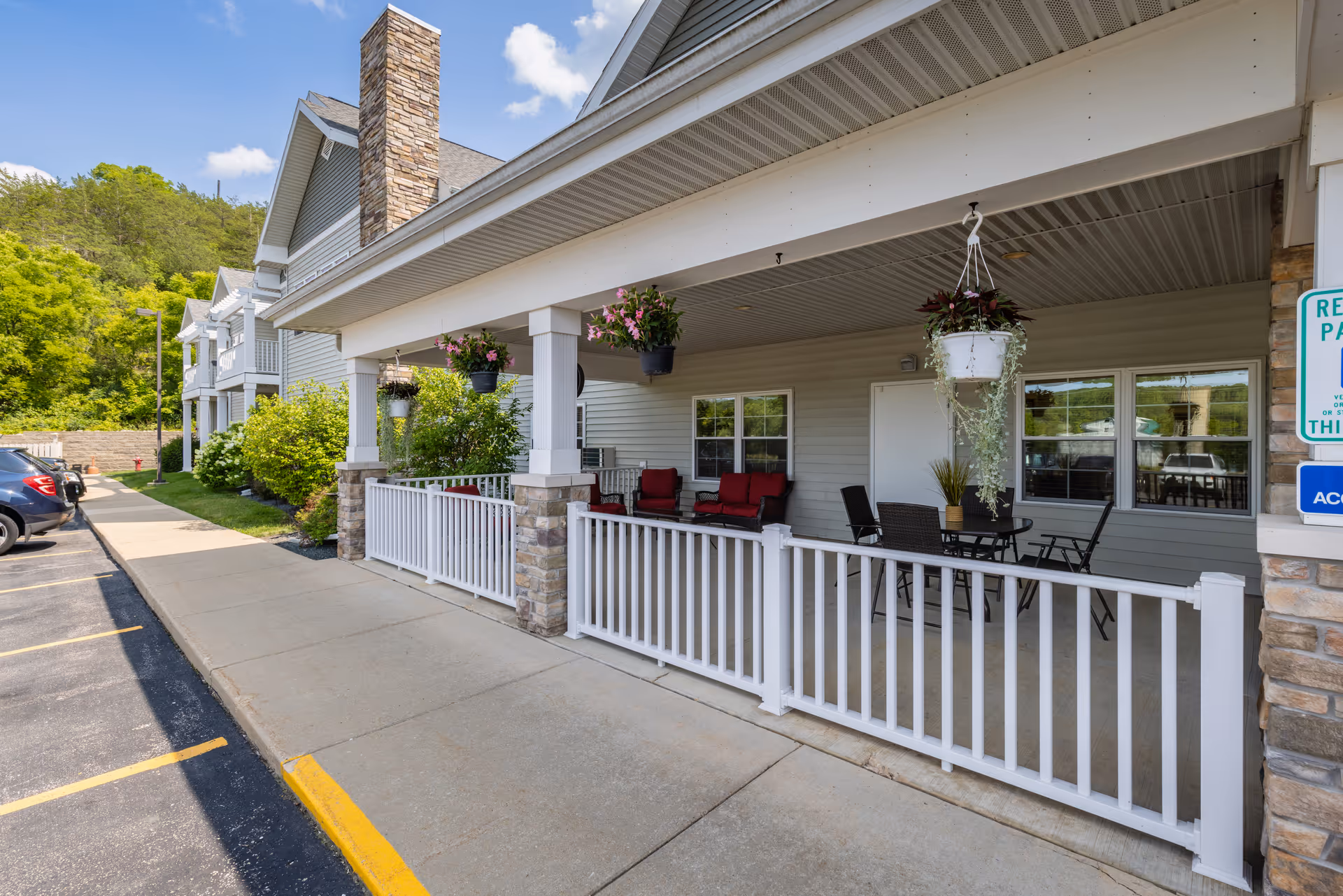 Covered front porch of a senior living facility with white railing, hanging plants, and outdoor seating next to a parking area.