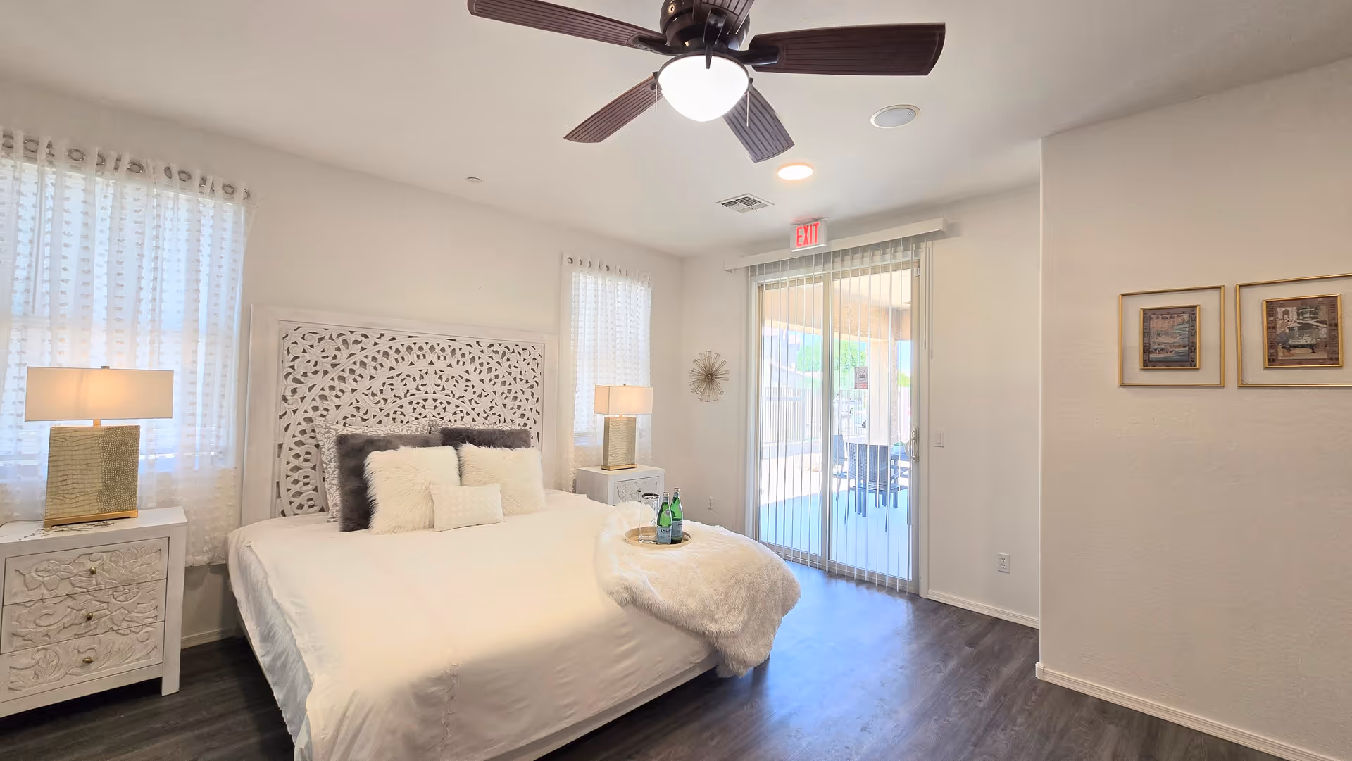 Bright bedroom with white decor, ornate headboard, nightstands, lamps, a ceiling fan, and sliding glass door.