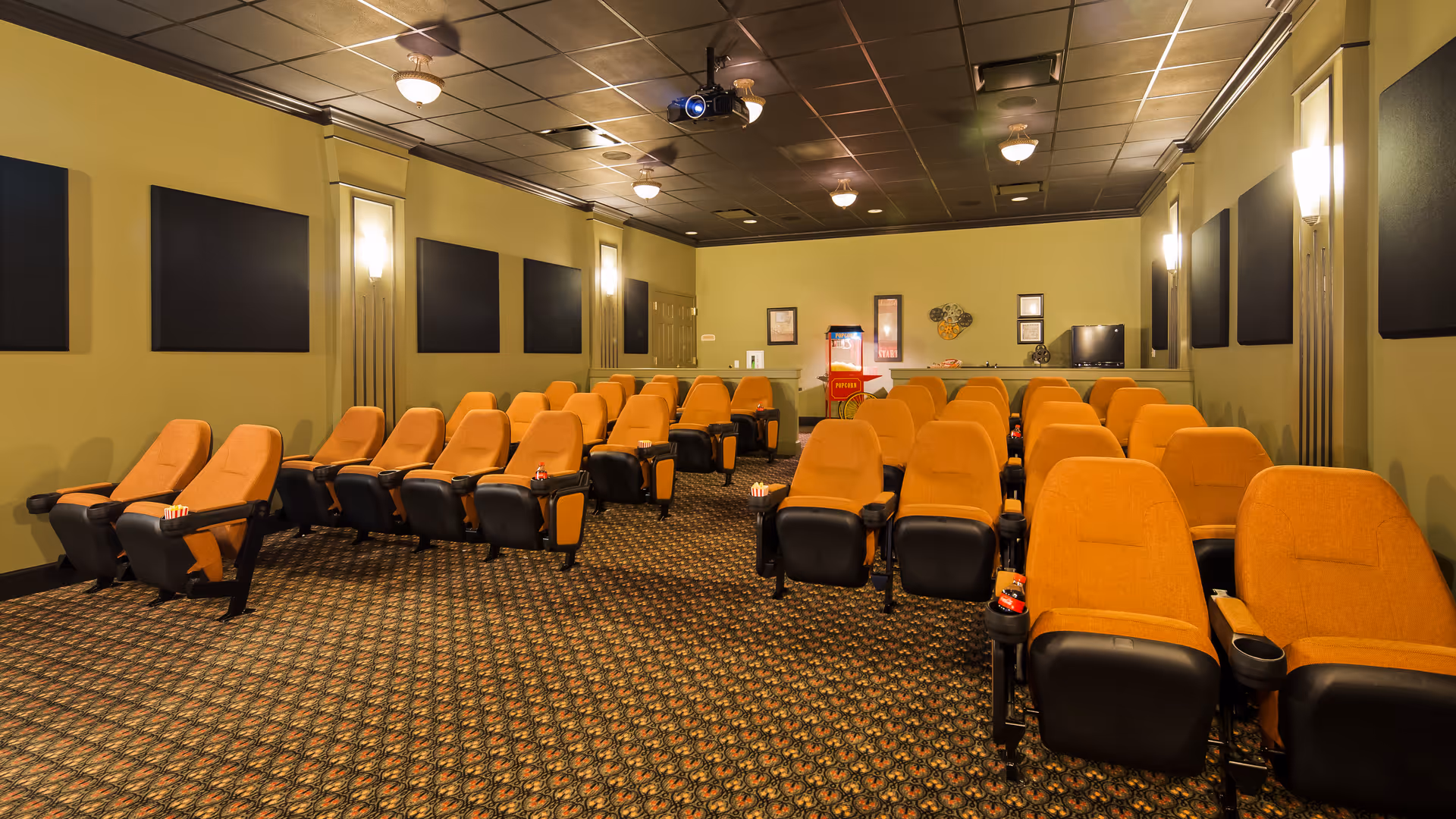 A small theater room with multiple rows of orange cushioned reclining seats arranged in front of a projector. The walls are painted green with black acoustic panels and wall sconces providing light. There is a popcorn machine and a small TV at the back of the room.