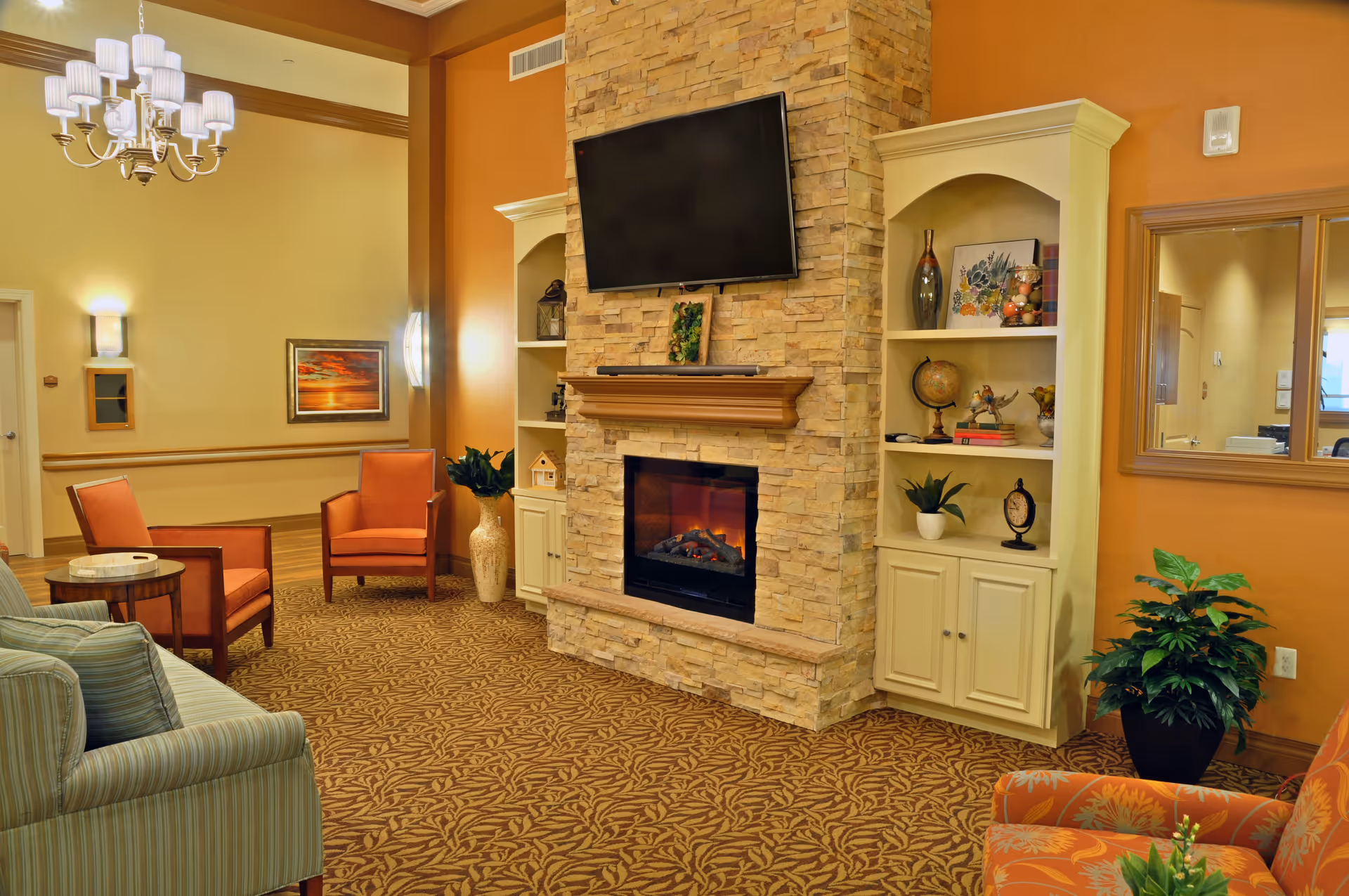 A cozy living room area with a stone fireplace and a mounted flat-screen TV above it. The room features built-in shelves on either side of the fireplace decorated with plants, books, and decorative items. There are several upholstered chairs in warm tones and a striped sofa, with a patterned carpet covering the floor. The walls are painted in warm colors, and a chandelier hangs from the ceiling.