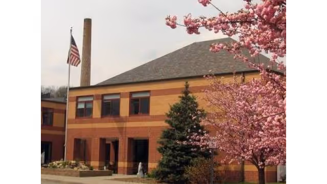 Two-story brick building with an American flagpole, a tall chimney, and pink flowering trees in front.