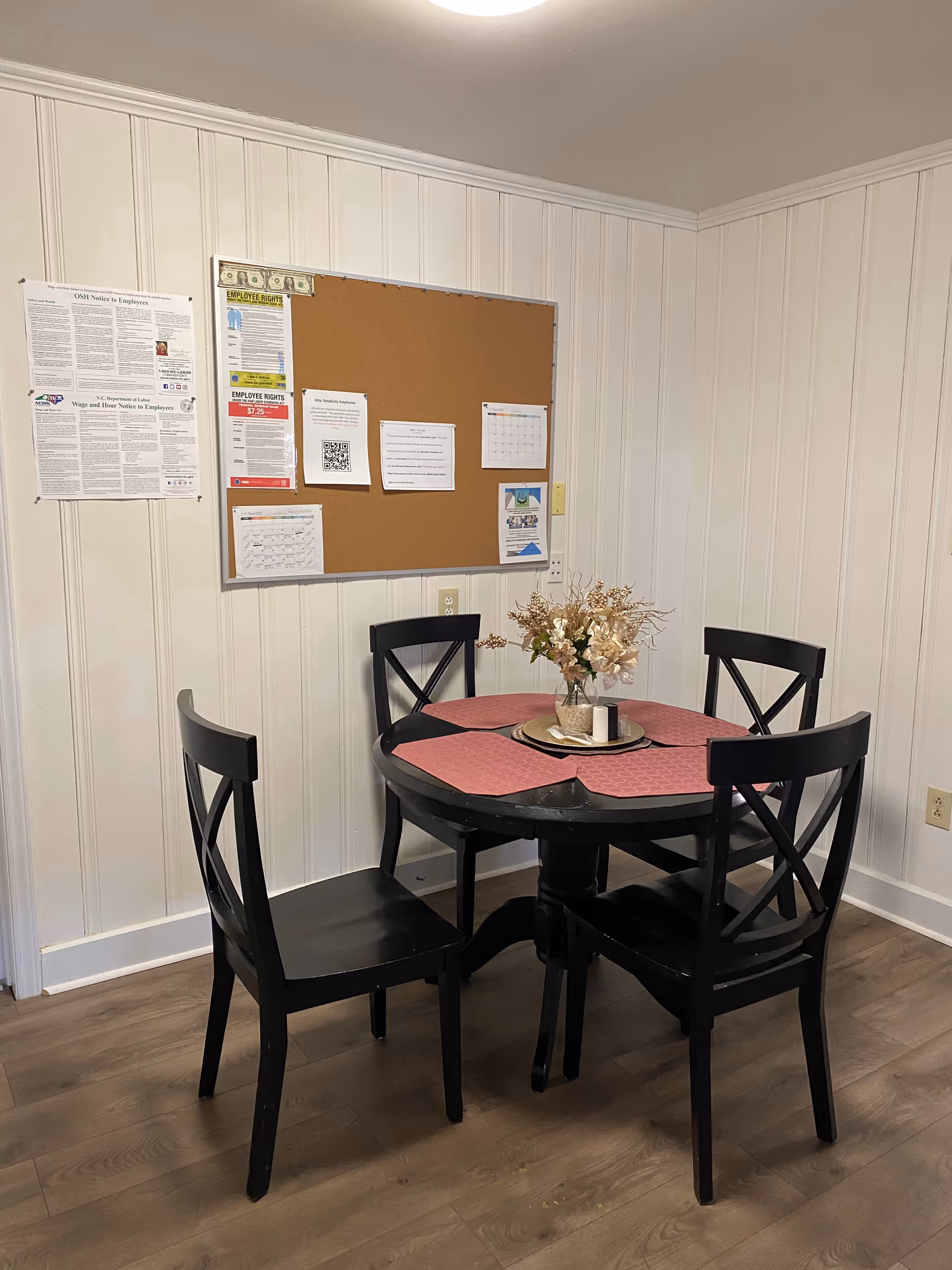Small dining area with round table, four black chairs, pink placemats, bulletin board.
