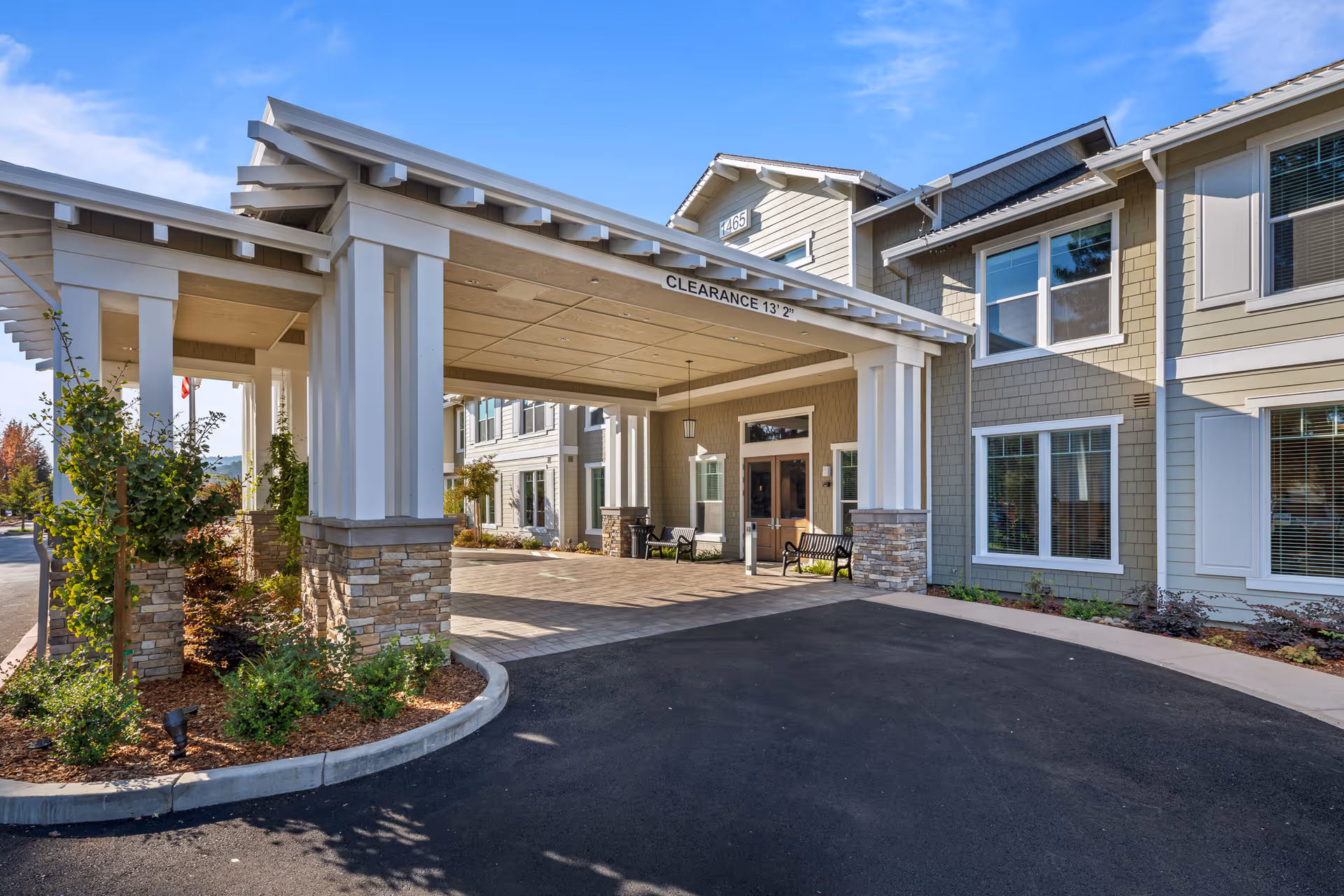 Entrance of a senior living facility named Oakmont of Lodi featuring a covered drop-off area with stone pillars and white beams, surrounded by landscaped plants and shrubs under a clear blue sky.
