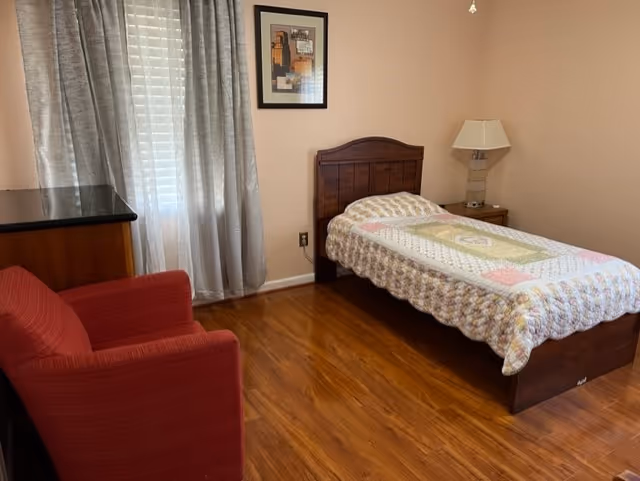 Furnished senior living bedroom with a wooden bed, nightstand, lamp, dresser, and red chair.