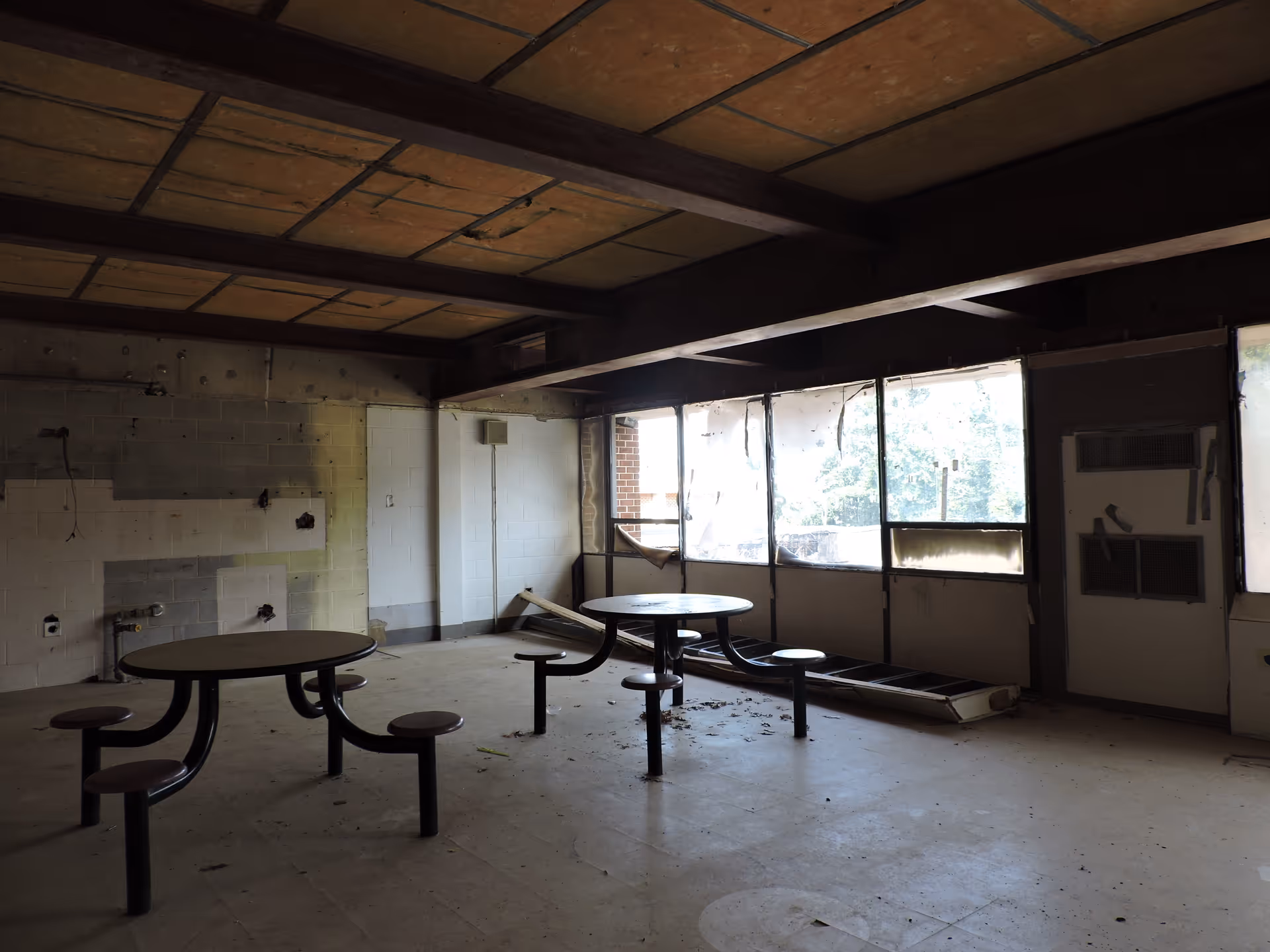Dilapidated indoor dining area with round tables, benches, and broken windows.