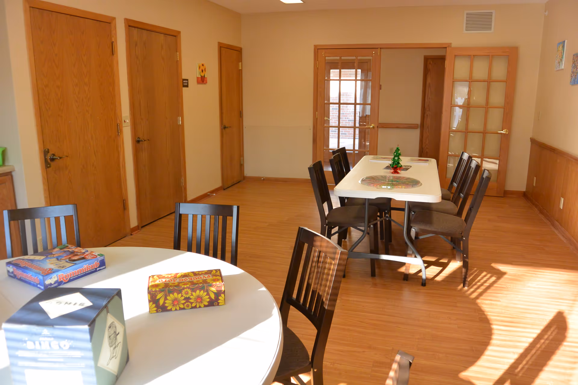 A well-lit room with wooden floors and beige walls featuring two tables with chairs around them. One table is round with board games on it, and the other is rectangular with a small decorative item in the center. There are wooden doors and glass-paneled doors along the walls.