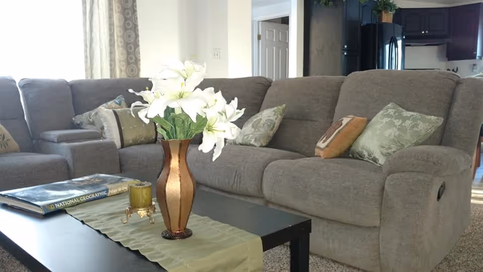 A cozy living room with a gray sectional sofa adorned with decorative pillows. In front of the sofa is a black coffee table with a green runner, a vase with white flowers, a candle holder, and a National Geographic book. The background shows part of a kitchen area with dark cabinets and a refrigerator.