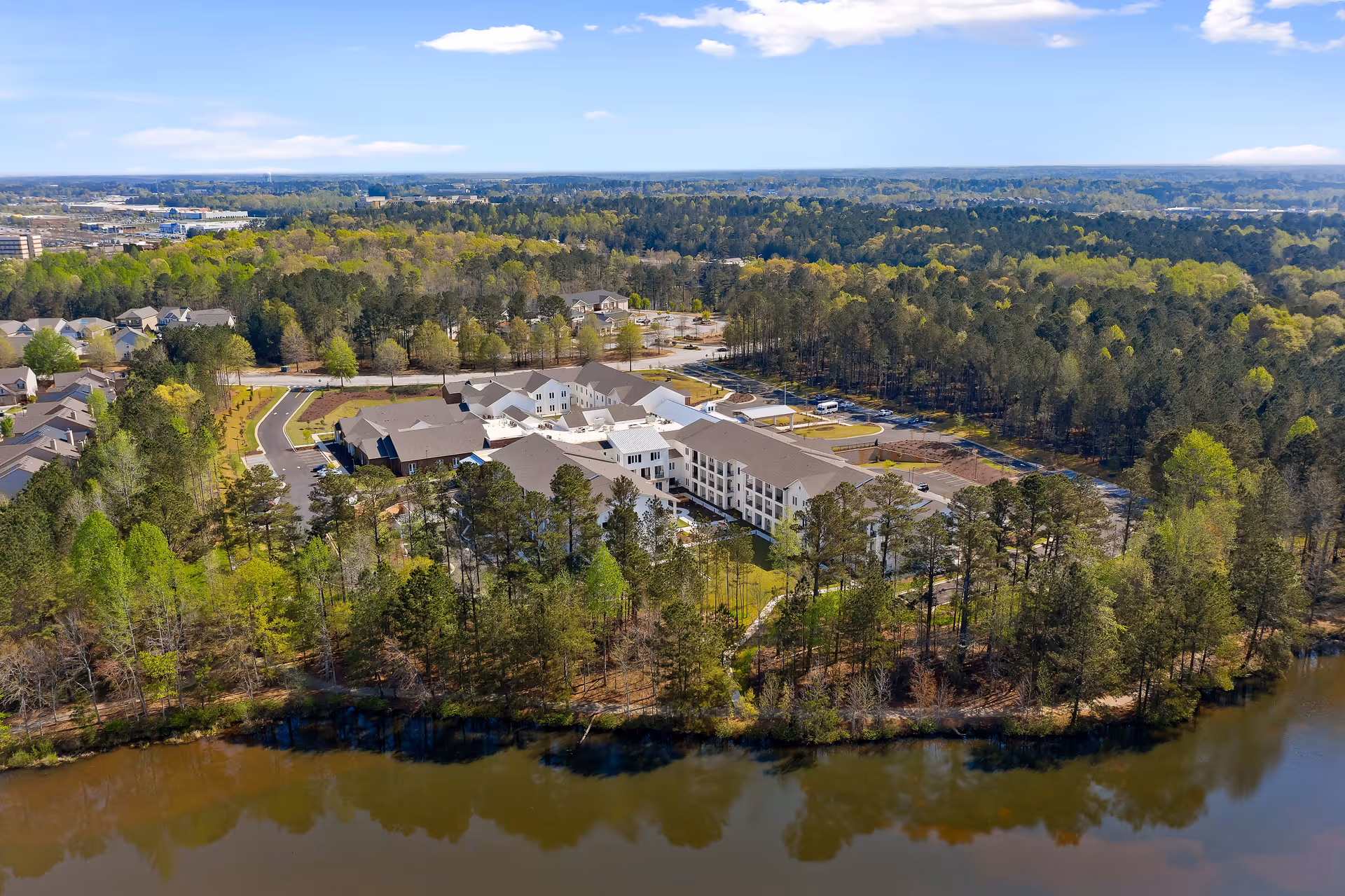 Aerial view of The Claiborne at Newnan Lakes senior living facility surrounded by trees and greenery, with a lake in the foreground and a parking area visible behind the building under a clear blue sky.