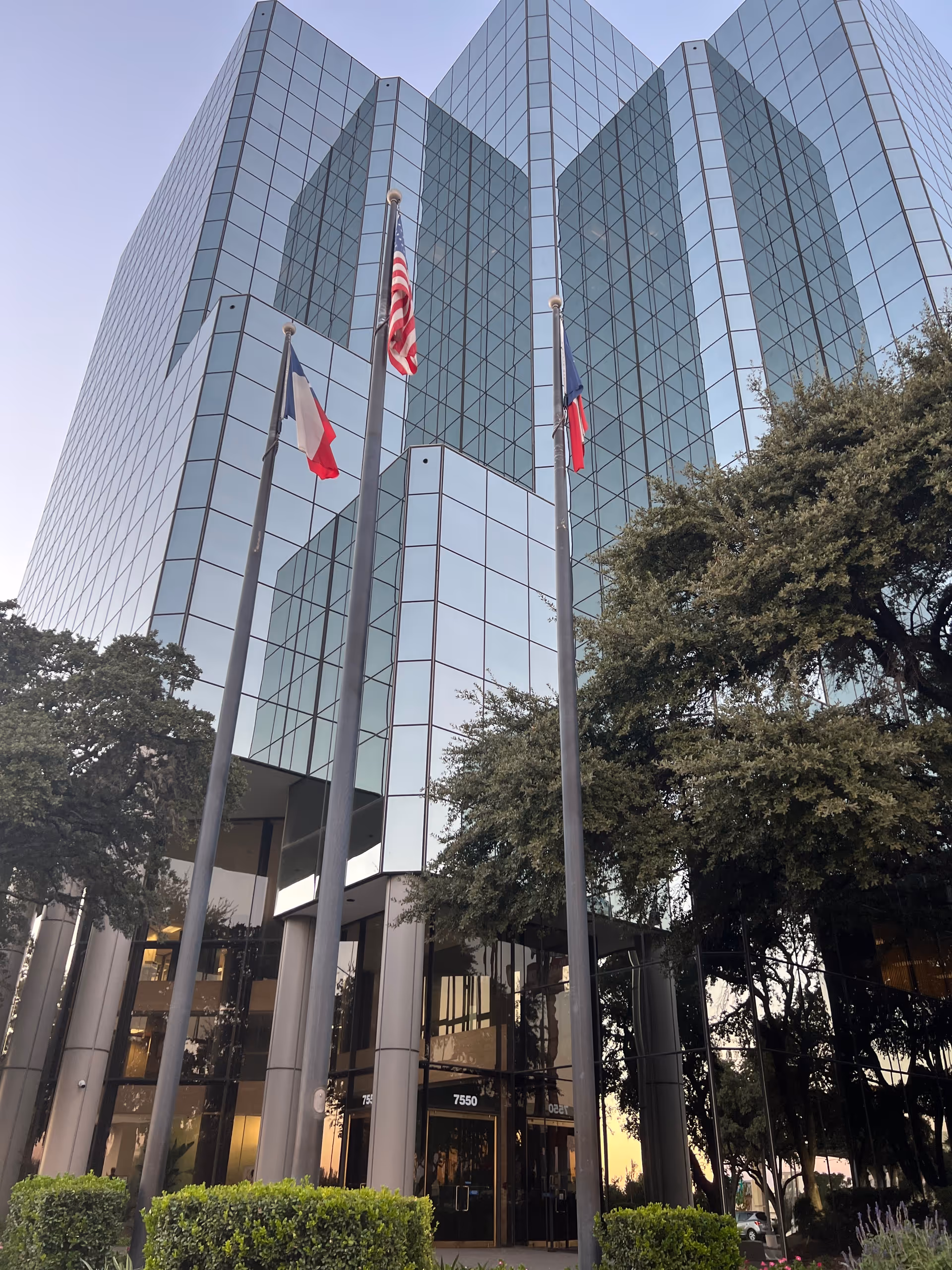 Tall glass front office building with flagpoles and surrounding trees.