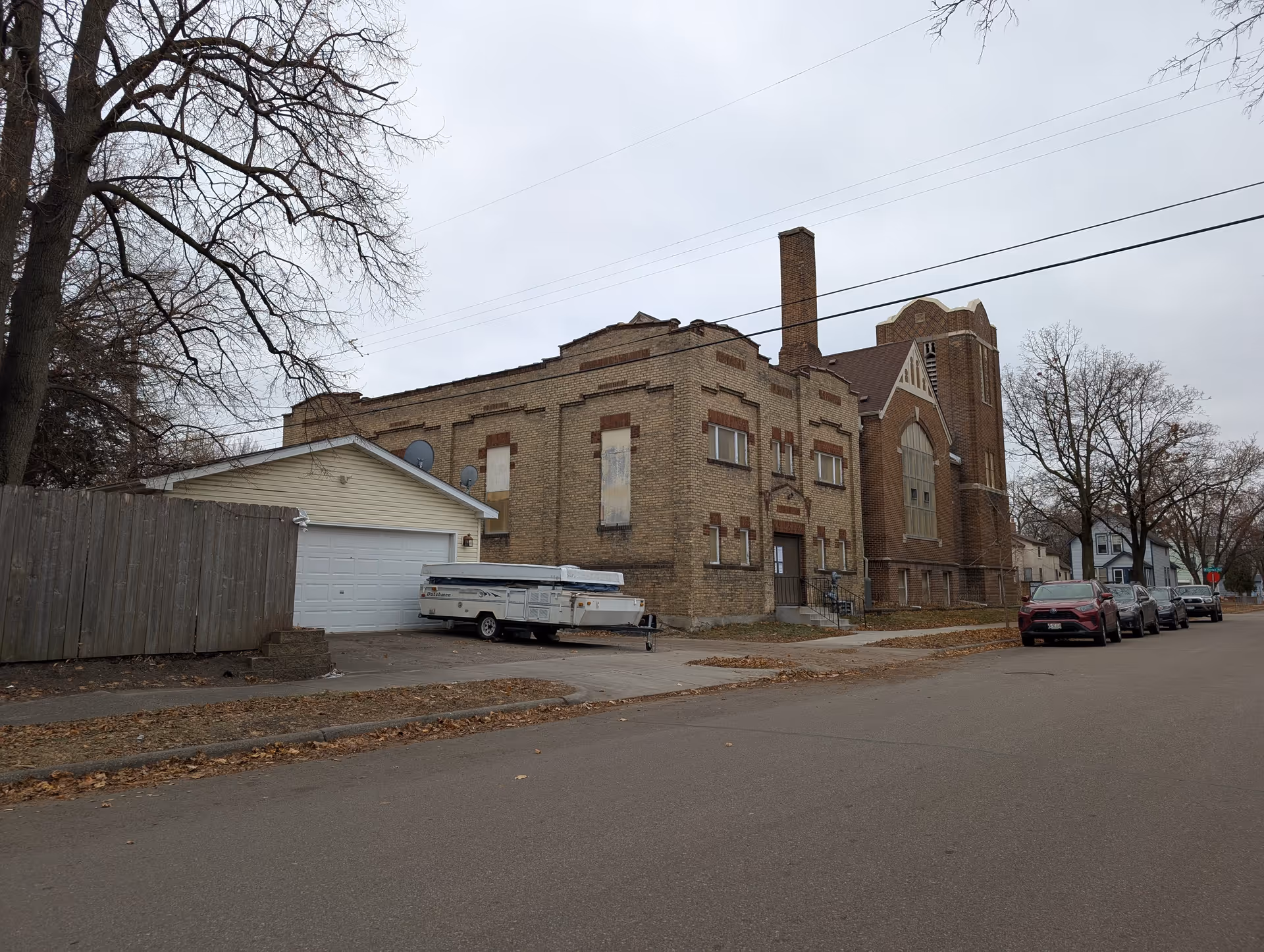 Large brick building with boarded windows and a tall tower on a residential street with parked cars and a garage in front.