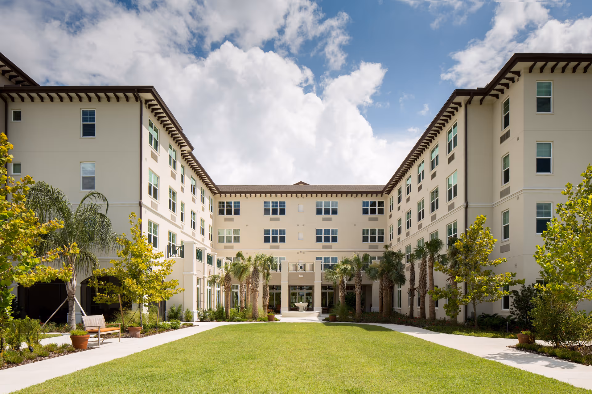U-shaped four-story senior living building surrounding a landscaped courtyard with lawn, palm trees, benches and walkways under a partly cloudy sky.