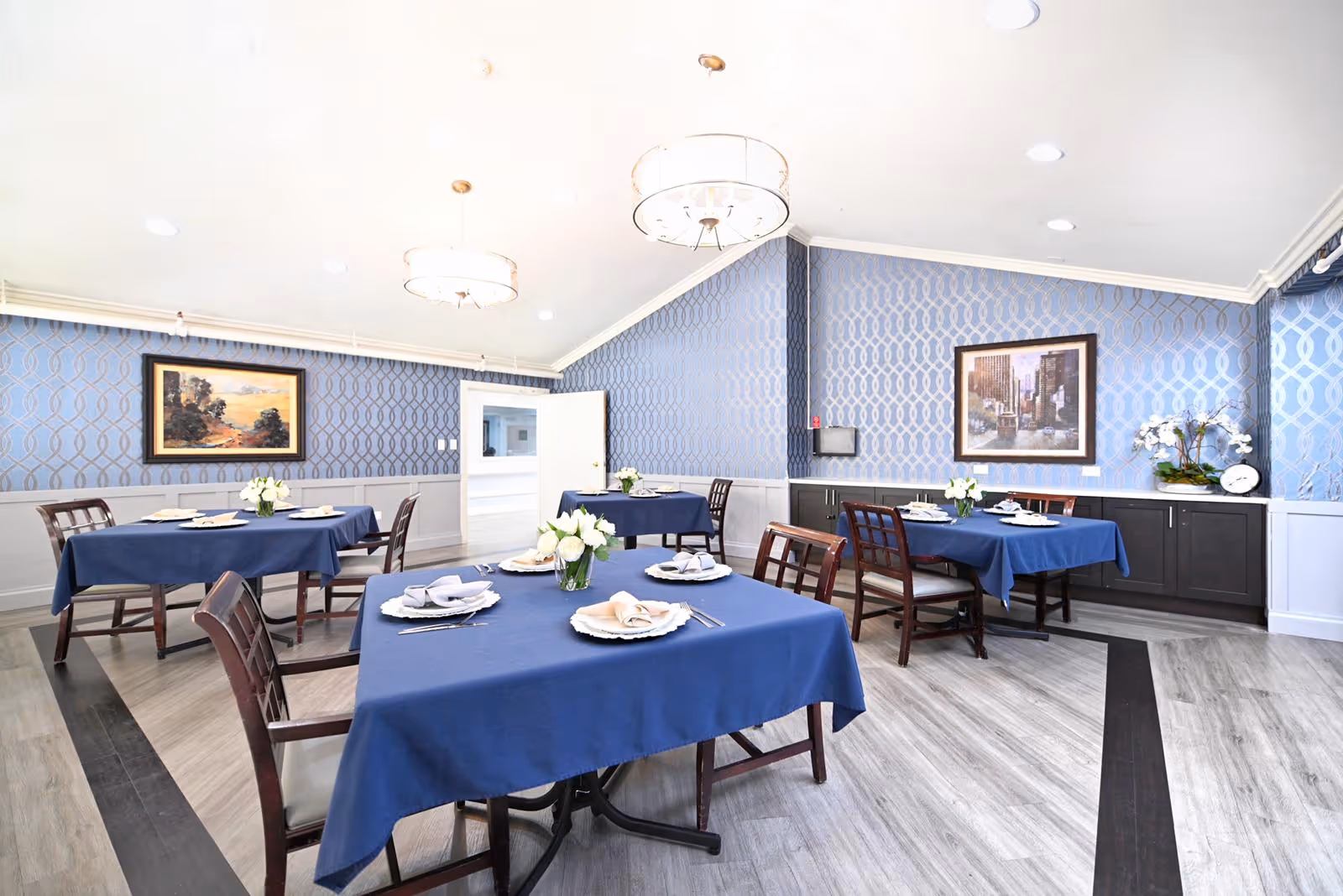 Bright dining room with multiple tables covered in blue tablecloths, wooden chairs, floral centerpieces, and patterned blue wallpaper.