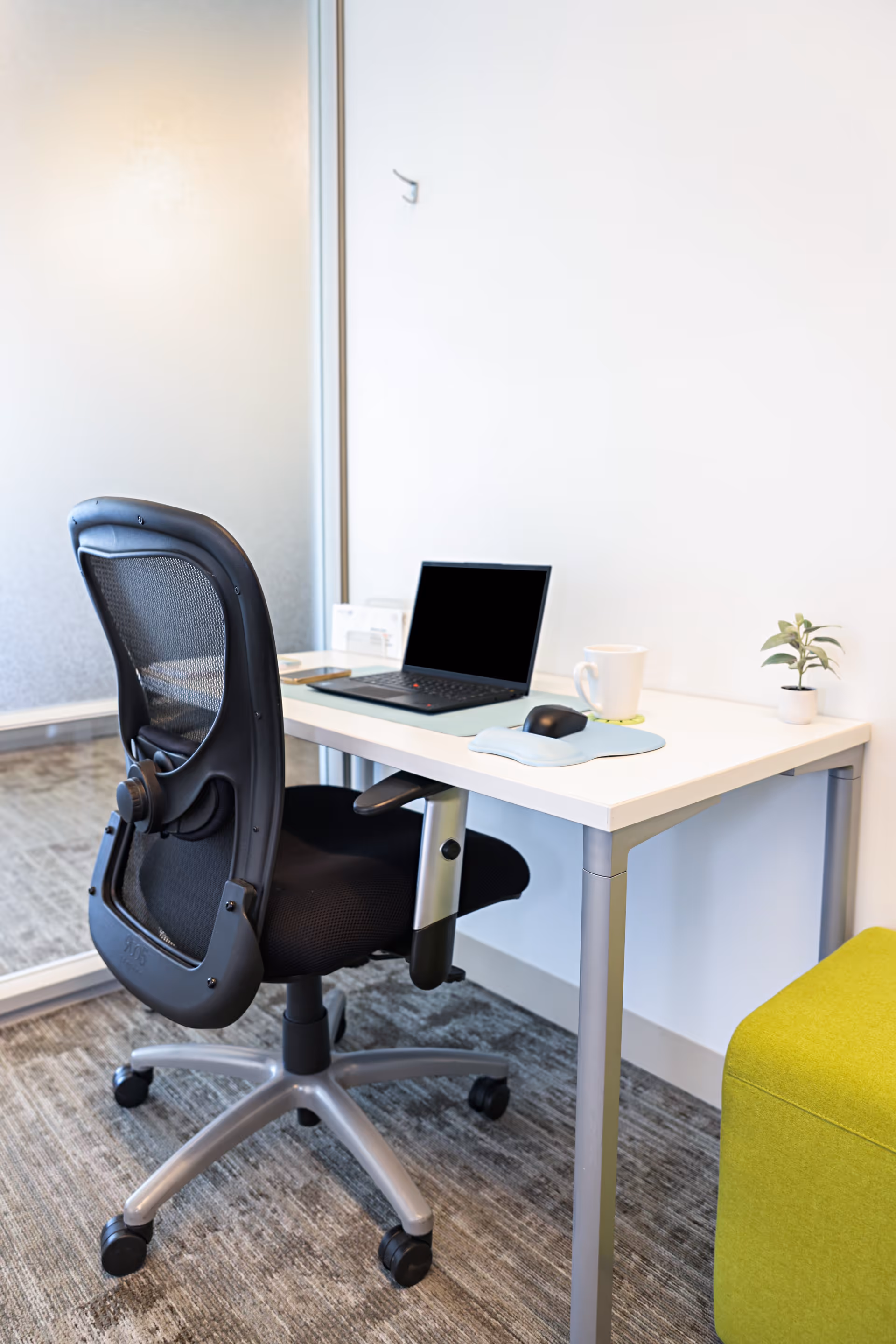 Modern office desk with laptop, mouse, mug, plant, and ergonomic chair indoors.