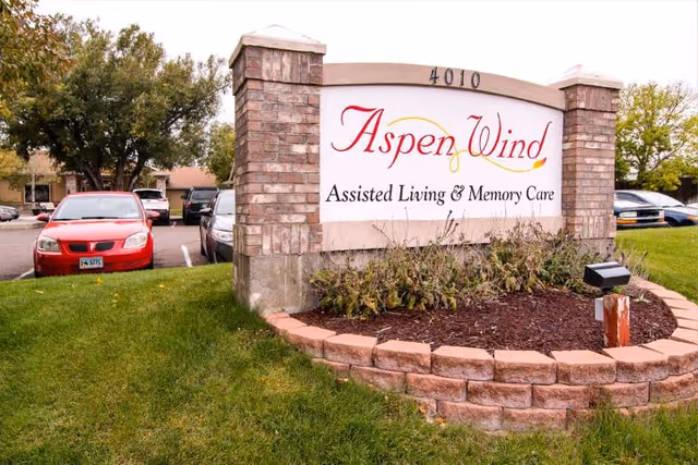 Outdoor view of a large sign for Aspen Wind Assisted Living & Memory Care, set in a landscaped area with brick pillars and a curved brick border. Several parked cars and trees are visible in the background.