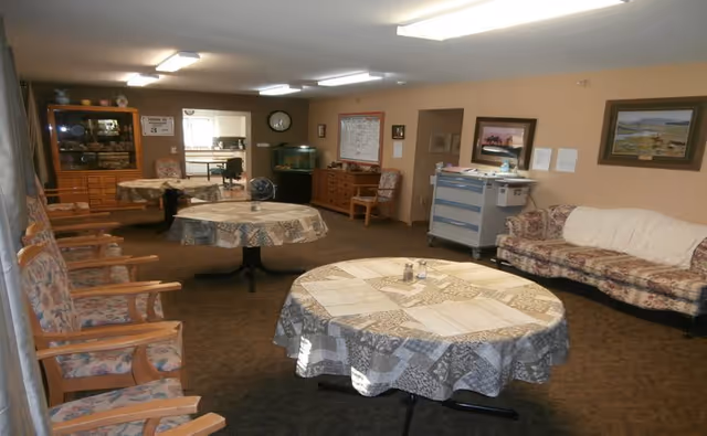 Indoor senior living facility lounge with round tables, patterned cloths, and cushioned seating.