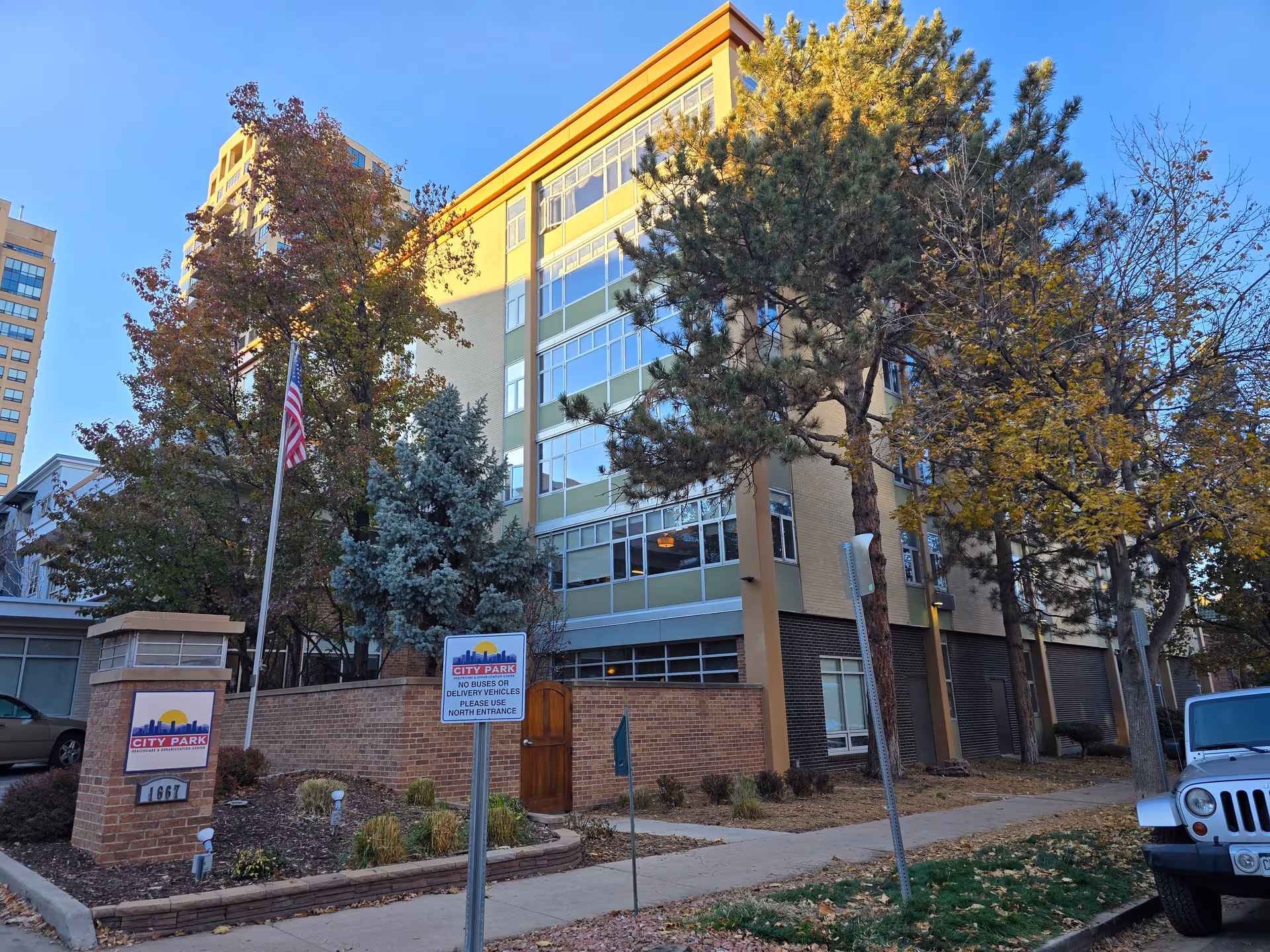 Exterior view of a multi-story residential building with large windows, surrounded by trees with autumn foliage. There is a brick sign at the front that reads 'CITY PARK Healthcare & Rehabilitation Center' and a flagpole with an American flag. A sidewalk and parked vehicles are visible in the foreground under a clear blue sky.