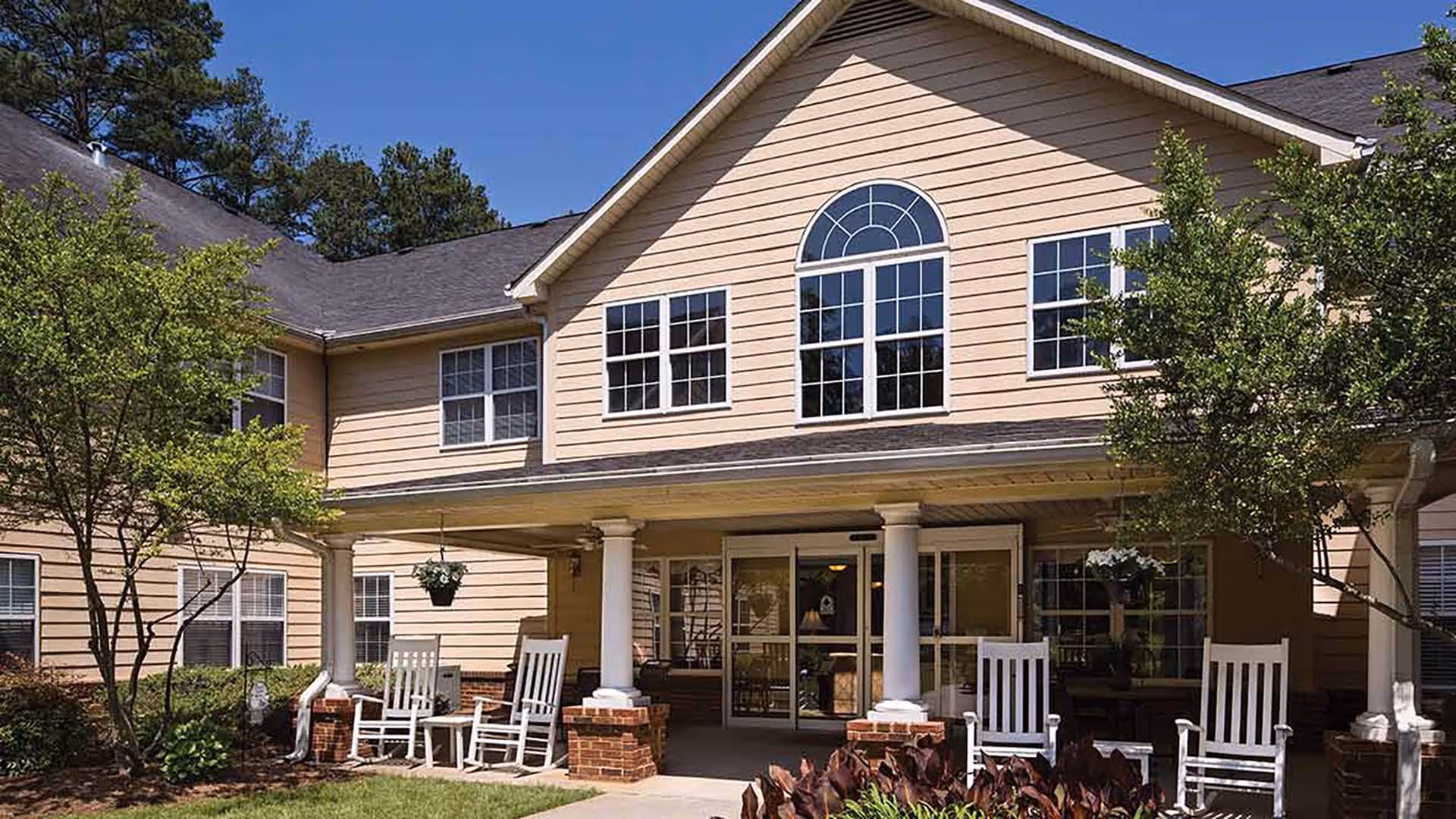 Exterior view of a senior living facility with beige siding, white columns, and large windows. The entrance has a covered porch with white rocking chairs and hanging flower pots. There are trees and landscaping around the building under a clear blue sky.