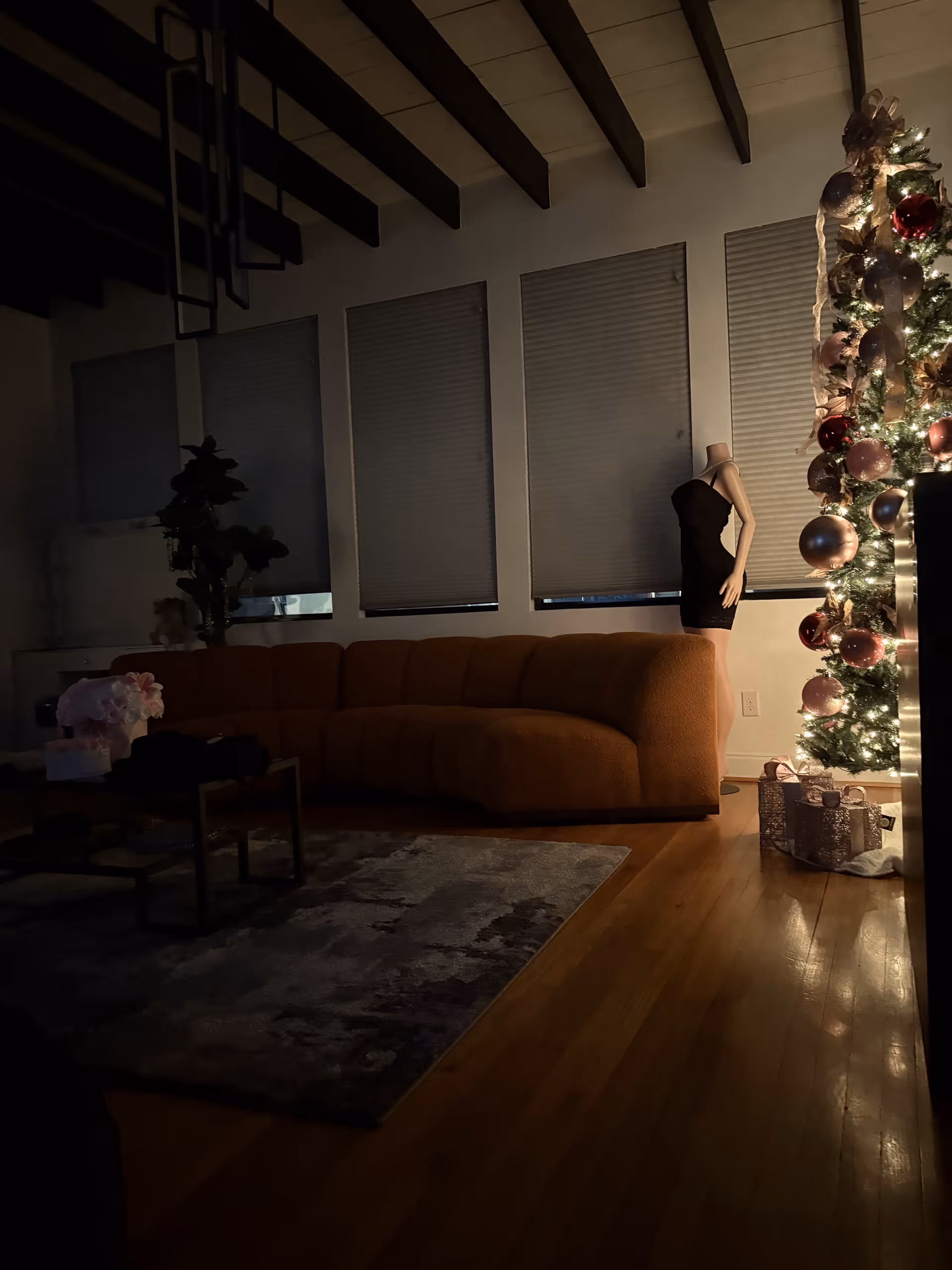 Dimly lit living room with brown sofa, rug, and a Christmas tree beside a mannequin.