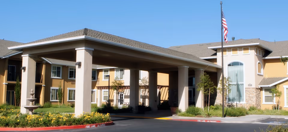 Exterior view of Prestige Assisted Living at Manteca showing the front entrance with a covered drop-off area, landscaped garden with a fountain, and an American flag on a flagpole against a clear blue sky.