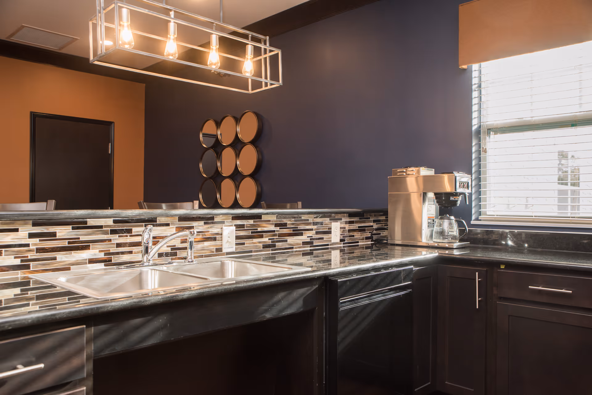 Modern kitchen area with a double stainless steel sink, a coffee maker on the counter, dark cabinetry, a mosaic tile backsplash, and a window with blinds letting in natural light. A decorative wall mirror with circular elements is visible on the dark blue wall.