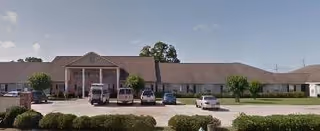 Front exterior view of a single-story senior living facility building with a covered entrance supported by columns, several parked cars in front, and some landscaping with bushes and trees.
