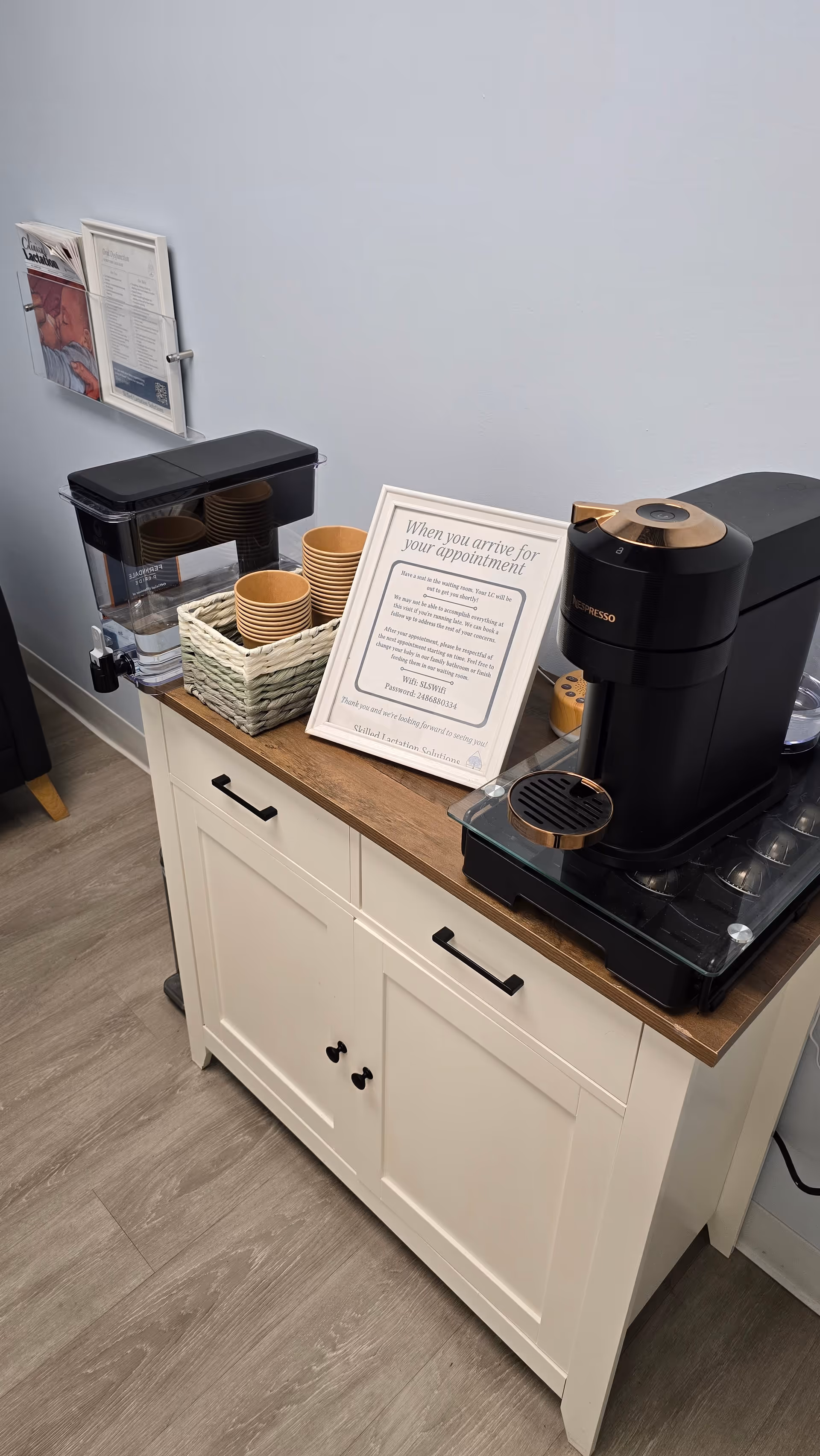 Interior coffee station with espresso machine, disposable cups, and framed sign on a white cabinet.